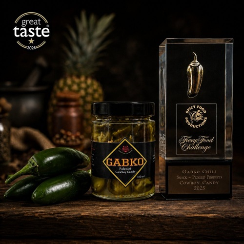 Jar of Gabko Cowboy Candy pickles beside fresh jalapeño peppers and a glass Fiery Foods Challenge award, with a pineapple in the background on a wooden surface.